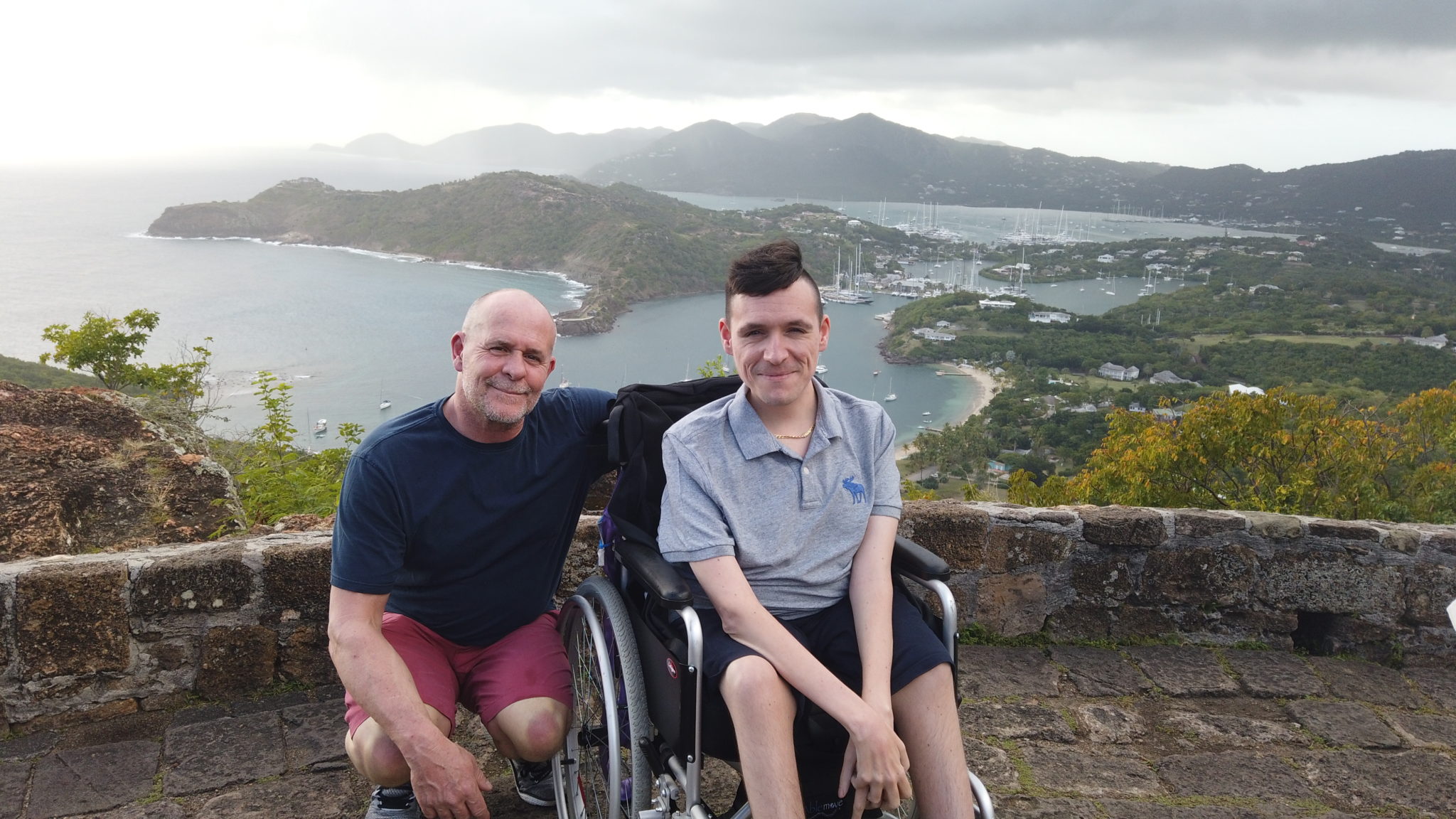 Josh's father crouched with his arm around Josh, who is sat in his manual wheelchair. Both facing the camera. In the background is Shirley Heights, with mountains, sea and harbour in the background with grey sky.
